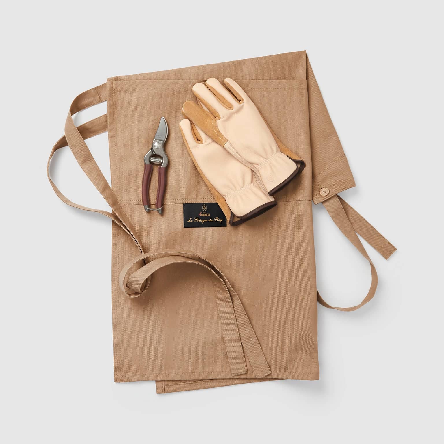 Tablier Le Potager du Roy x Le Laboureur plié, avec une paire de gants et un sécateur posés sur la toile brune.