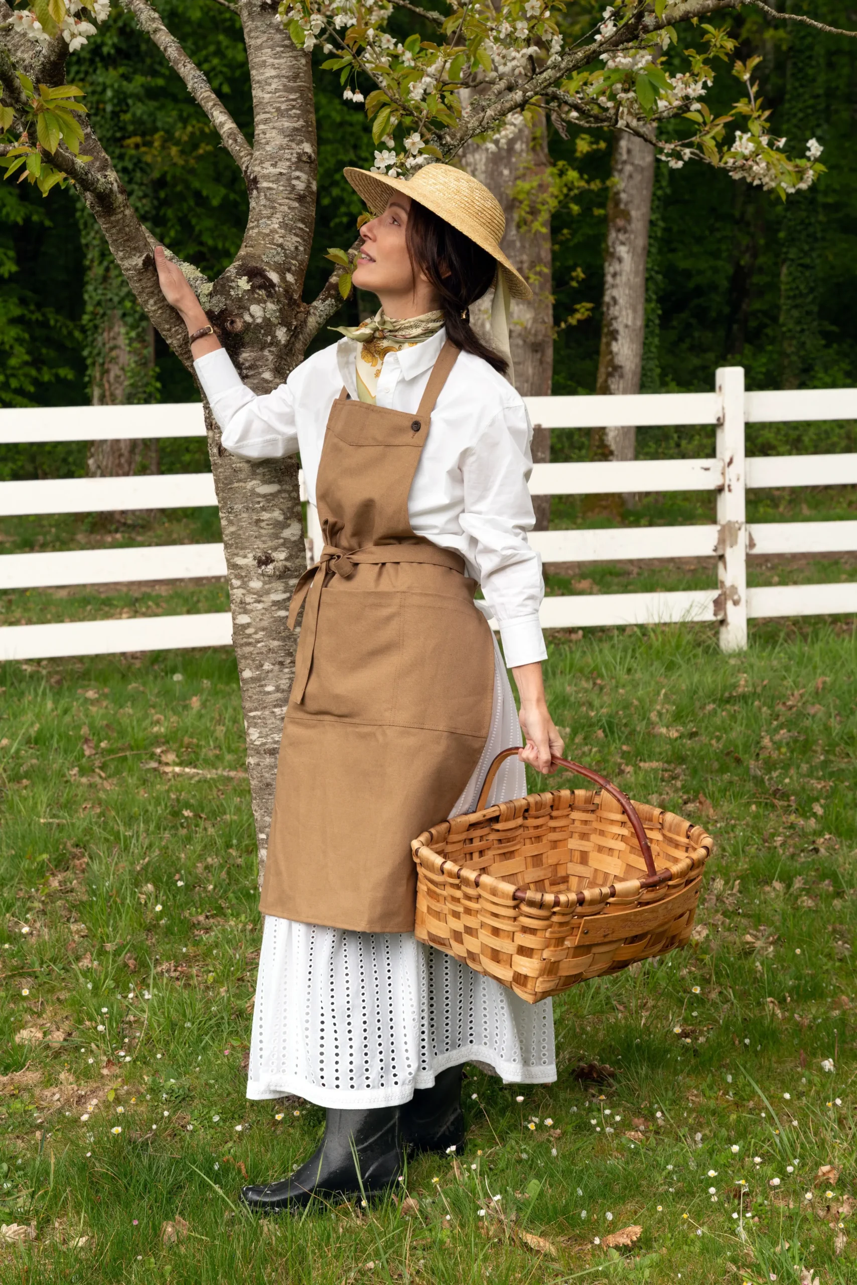 Tablier brun Le Potager du Roy x Le Laboureur porté dans un jardin, avec une ceinture nouée à la taille et un panier à la main.
